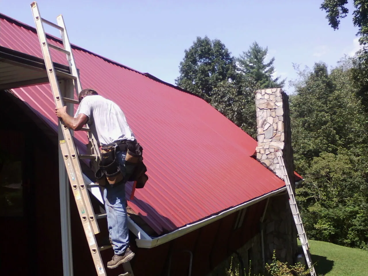 Roofer on a ladder beside a red metal roof and stone chimney — Soffit & Fascia Repair by Roofing Brothers in West Sunbury